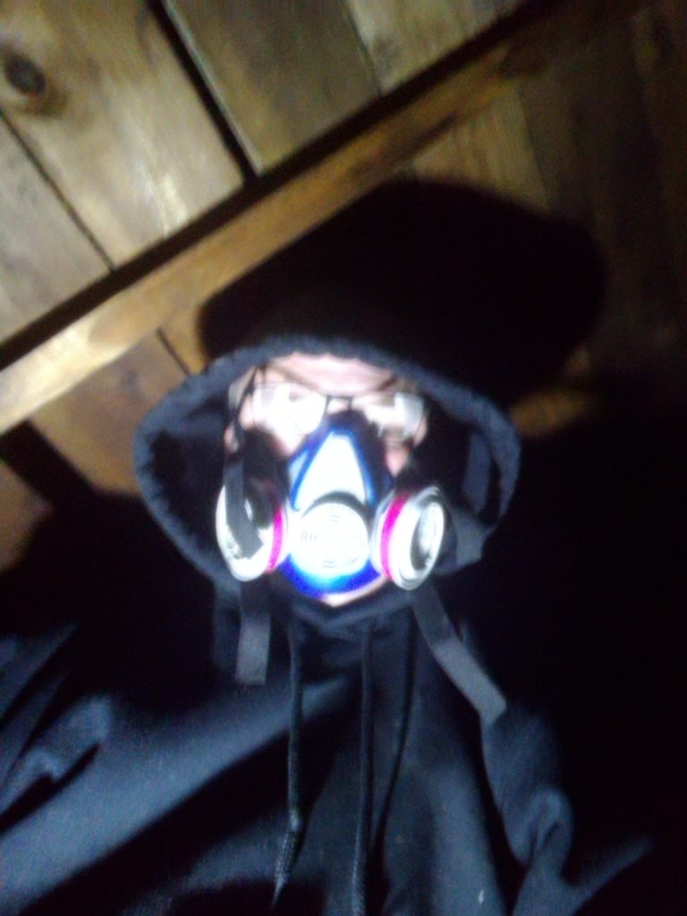 A blurry selfie of me hearing a black hooded sweatshirt and a dust mask while working in the attic. Planks for the rook are just behind me.