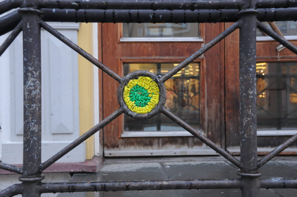 A piece of yellow and green crochet is set into a metal railing. In the background is a wooden door that leads into the building.