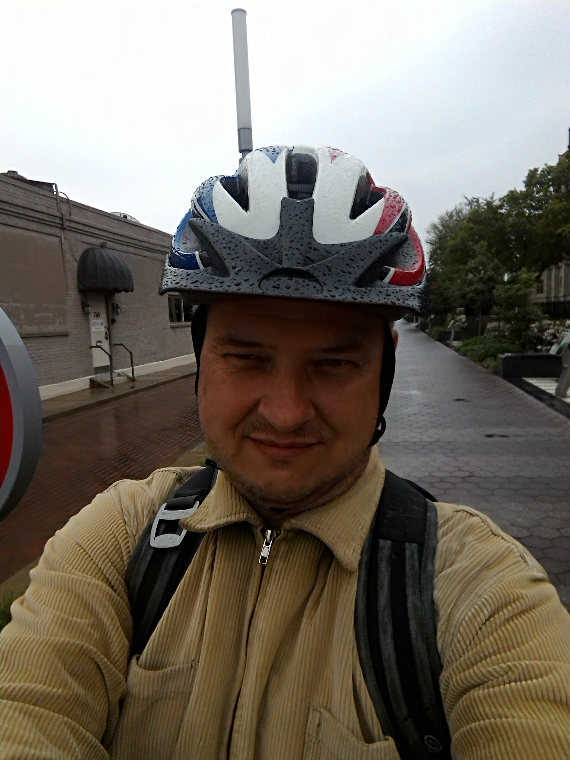 I'm wearing a green corduroy jacket, a backpack, and a rain-spattered bicycle helmet. In the background is a building, some trees, and the brick-paved trail.