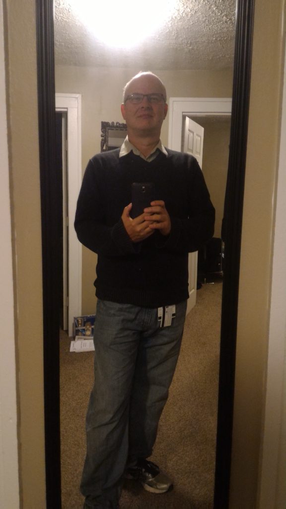 This photo shows me wearing running shoes, baggy jeans, and a black sweater over a collared shirt. An ID badge hangs from my belt and is visible just below the hem of the sweateer. Thebackground shows the black frame of the mirror against a beige wall.