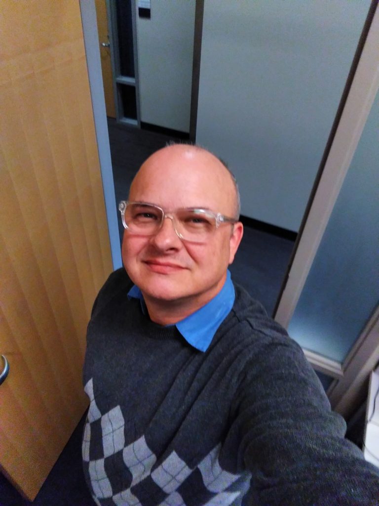 In the photo, I'm wearing a grey Argyle sweater over a blue-collared shirt My face is clean shave and adorned with clear-framed eyeglasses. Behind me is an open door and wall on the other side of the hallway. a wall .