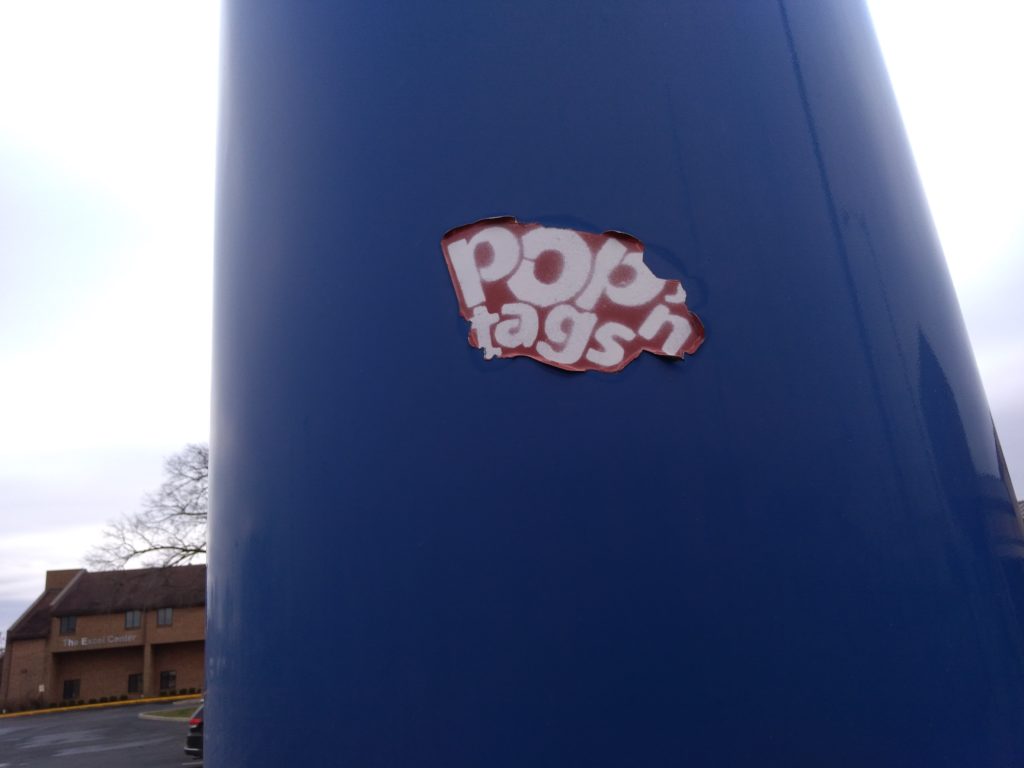 A decal reading Pop'n tags is stuck to an outdoor sign of some kind. The edges of the decal are peeling away, indicating age.
