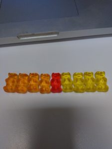 A red gummi bear sits in the middle of a line of yellow and orange gummi bears.