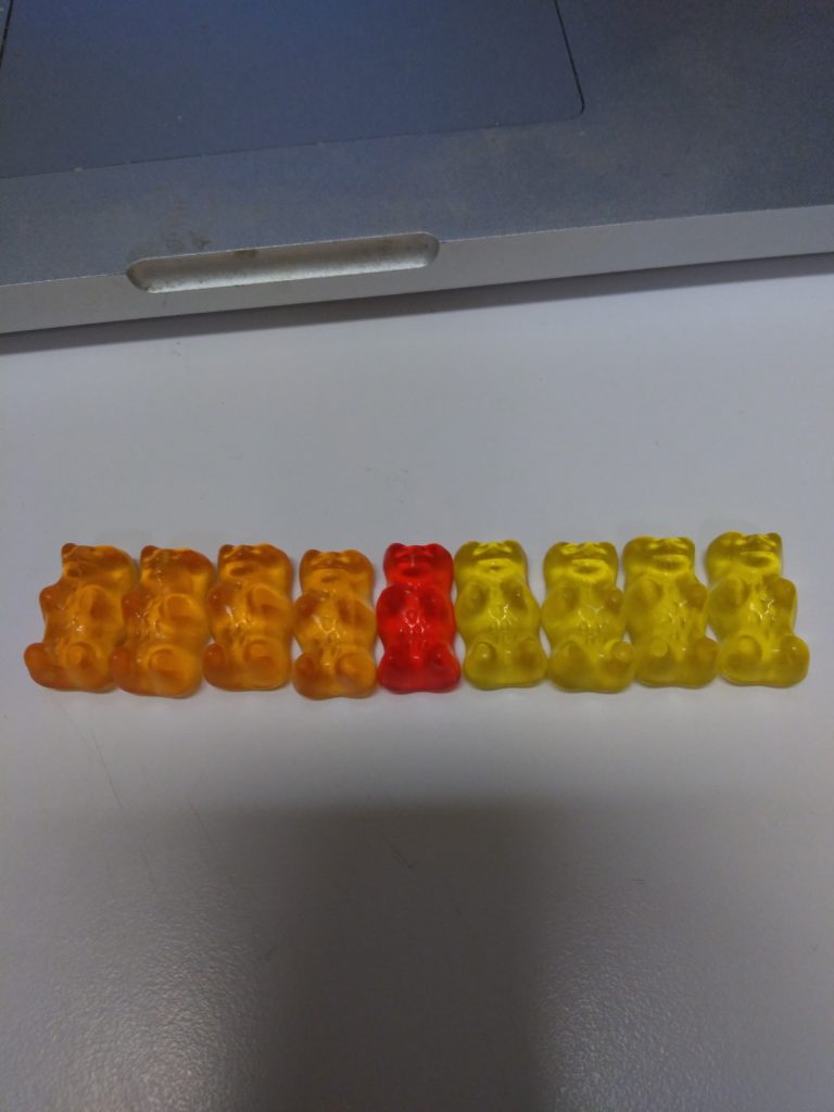 A red gummi bear sits in the middle of a line of yellow and orange gummi bears.