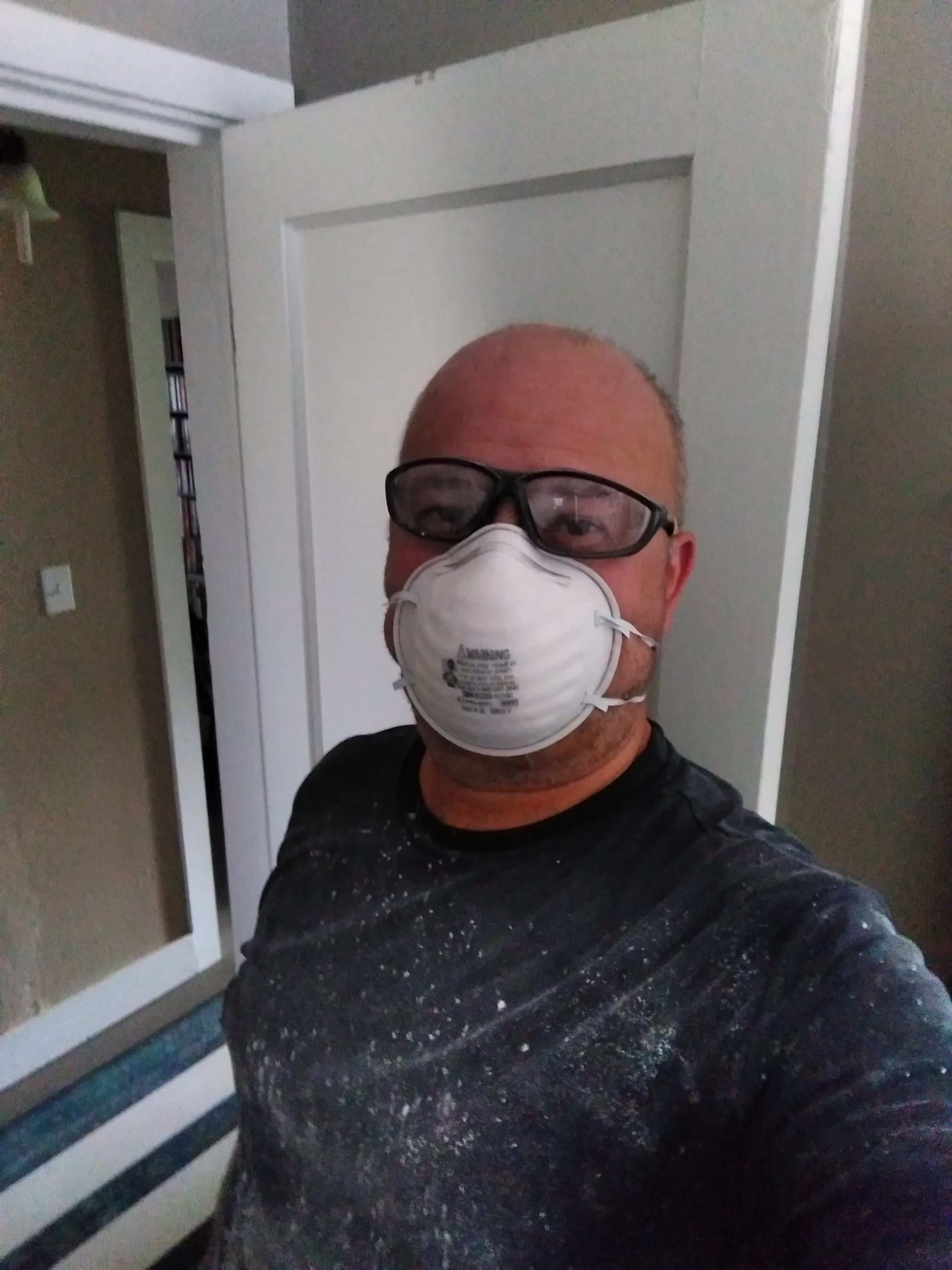 I'm wearing a dark shirt covered in plaster dust, an N95 face mask, and safety glasses. In the backgrounds is an open wodden door and the beige painted walls of the hallway..