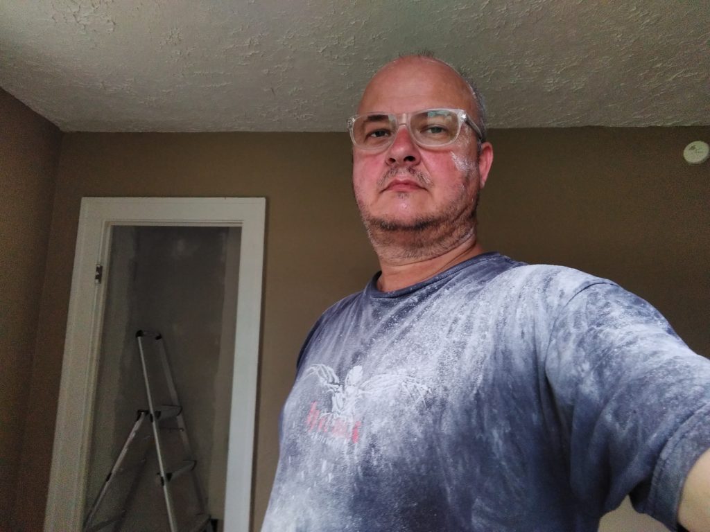 I', wearing a navy blue t-shirt covered in plaster dust, and clear-framed eyeglasses. My face diplays a mixture of stubble and plaster dust. The background shows the wall of the kitche and the doorway of the pantry.