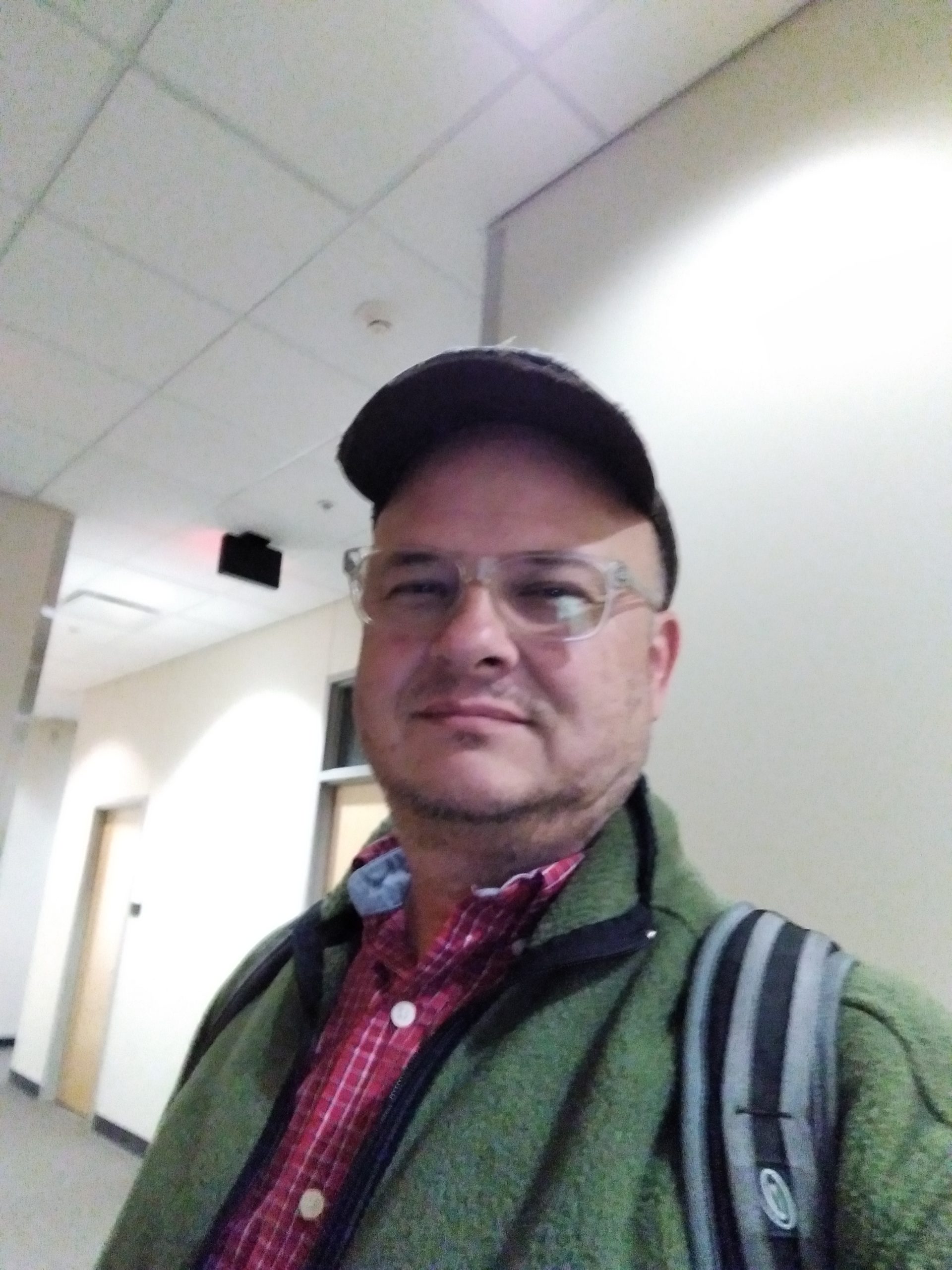 In this photo I'm wearing a green fleece jacket, a backpack, a red-checked collared shirt, clear-framed eyeglasses, and a well-worn baseball cap. In the background is the hallway with its beige-painted walls.