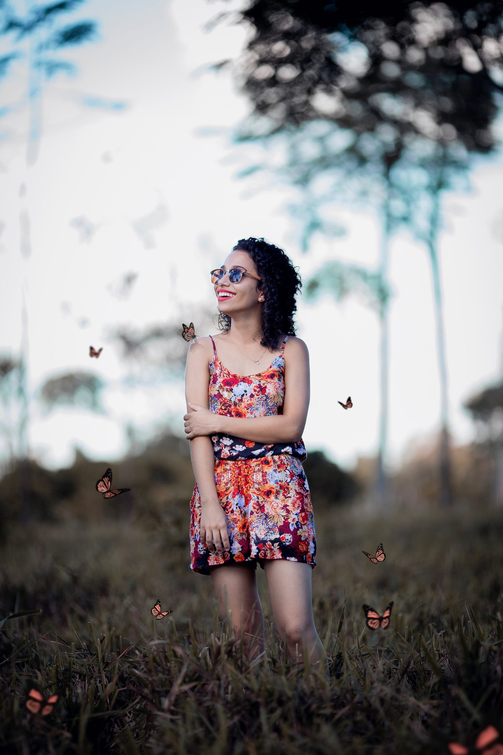A woman wearing sunglasses and a short dress is standing in a field and she is smiling as butterflies are flying around her.