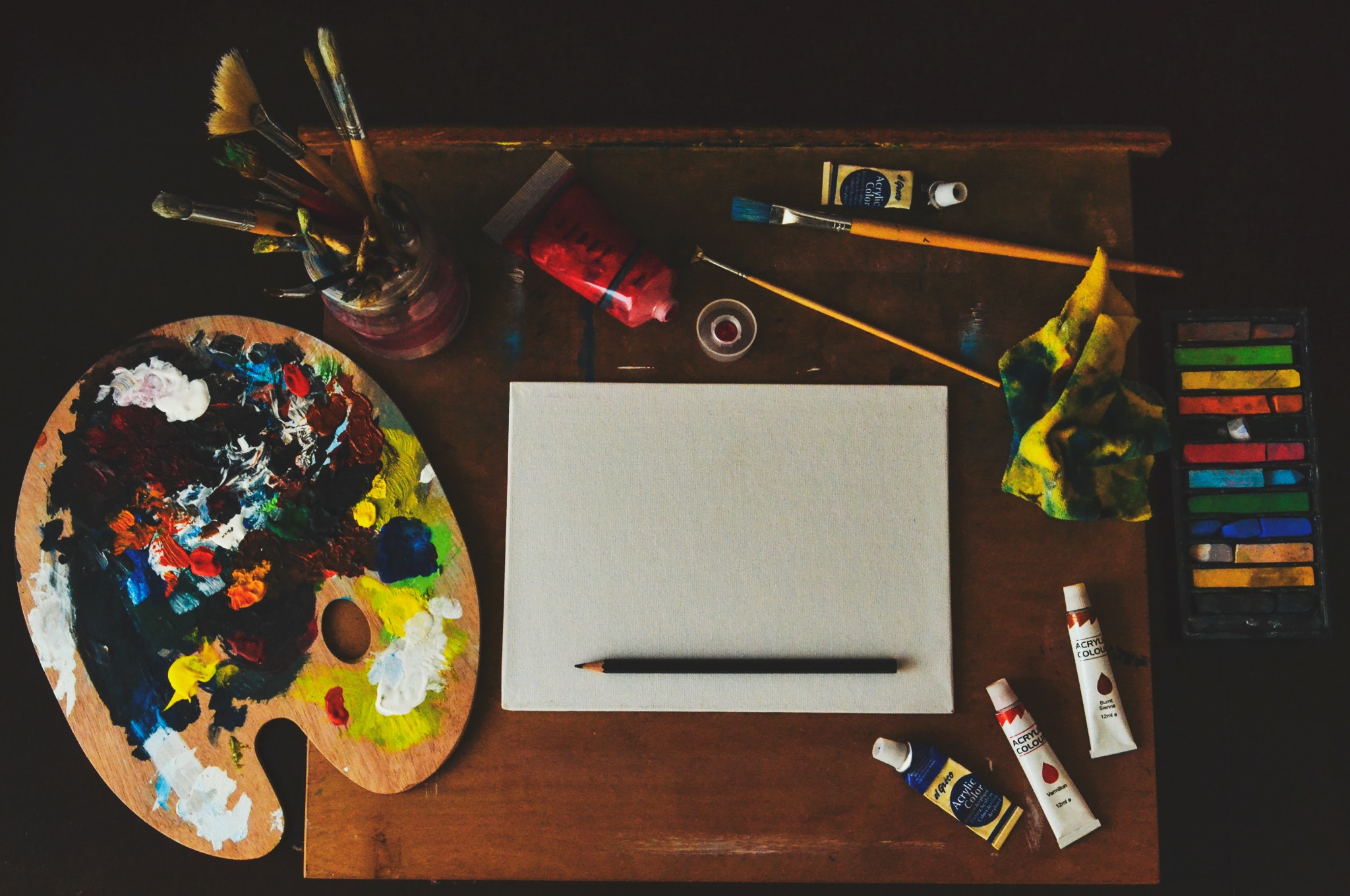 A color photograph showing an artist's canvas, tubes of paint, a tray of oil pastels, some used paintbrushes and palette.