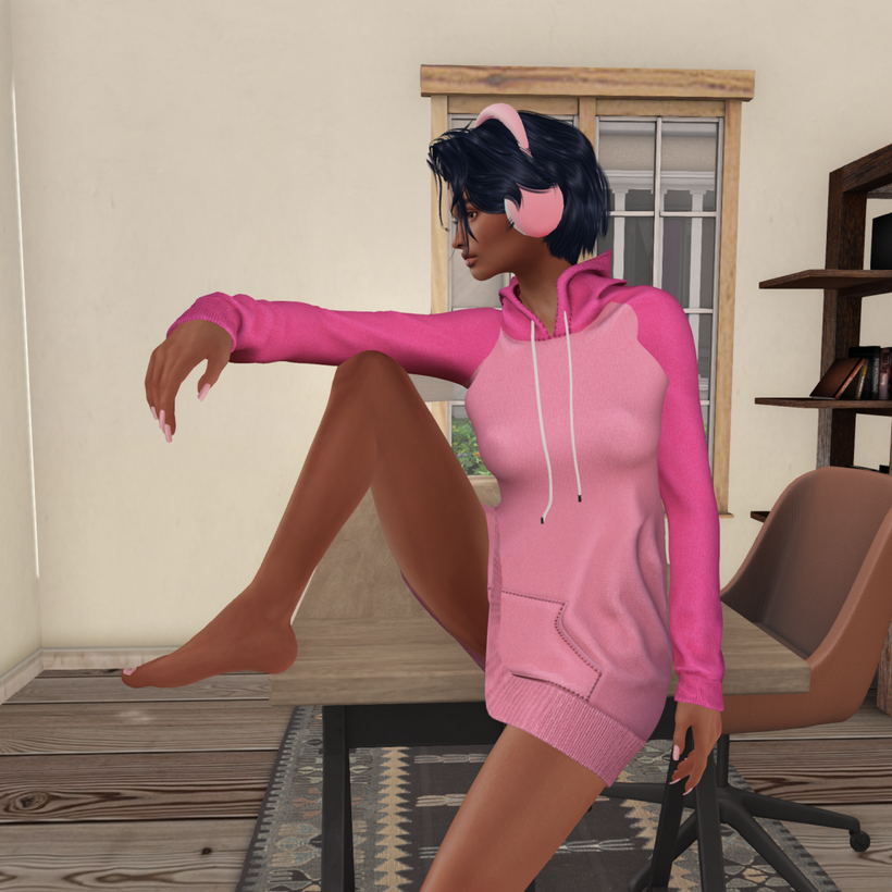 Kalyani is dressed in a pink hoodie and wearing headphones while sitting on a desk with her right leg up on the desk and her right arm stretched out over the knee.
